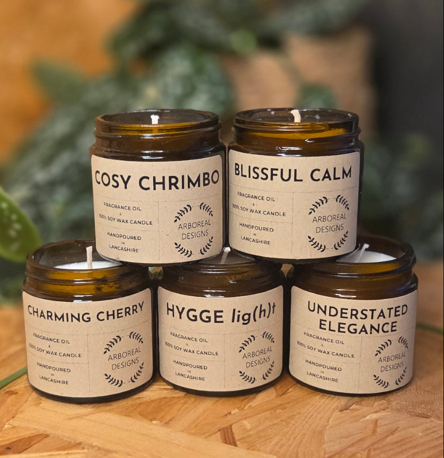Jar of various scented soy wax candles with labels on a wooden surface and green plant background