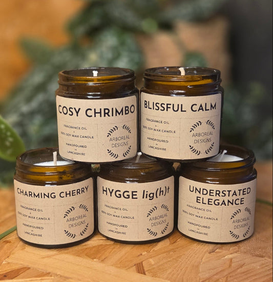 Jar of various scented soy wax candles with labels on a wooden surface and green plant background